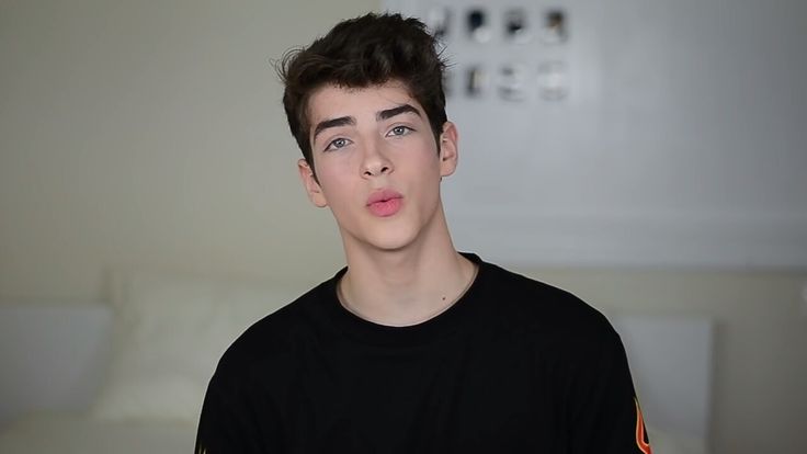 A close-up, smiling portrait of Manu Ríos, showcasing his signature cute and stylish aesthetic. He is looking directly at the camera with his striking green eyes, against a neutral background that highlights his sharp features and modern fashion sense.