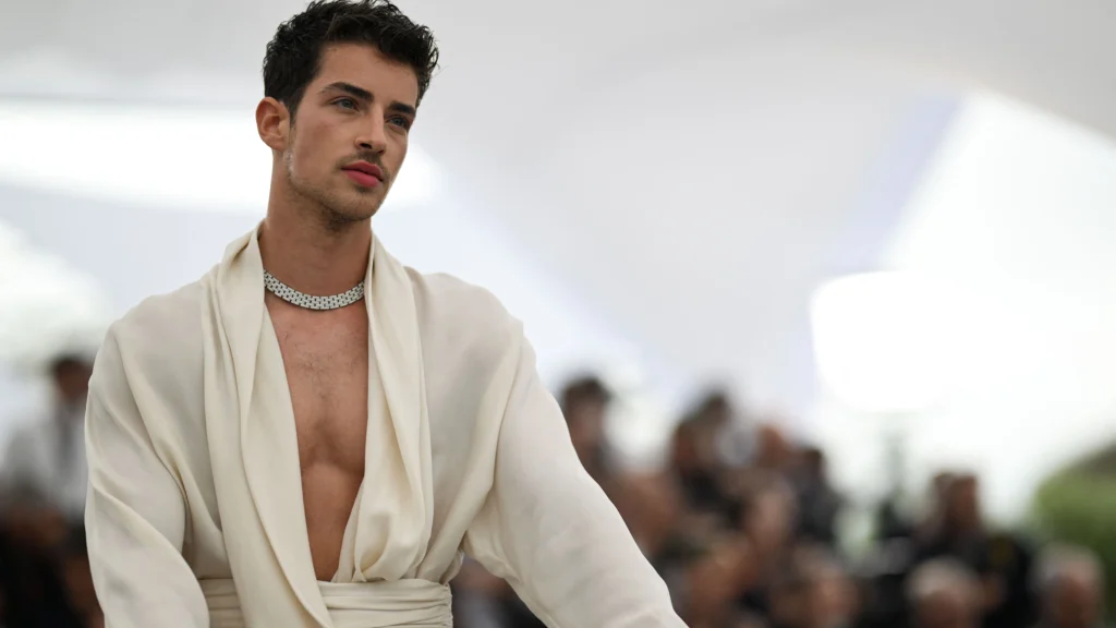 A close-up, smiling portrait of Manu Ríos, showcasing his signature cute and stylish aesthetic. He is looking directly at the camera with his striking green eyes, against a neutral background that highlights his sharp features and modern fashion sense.